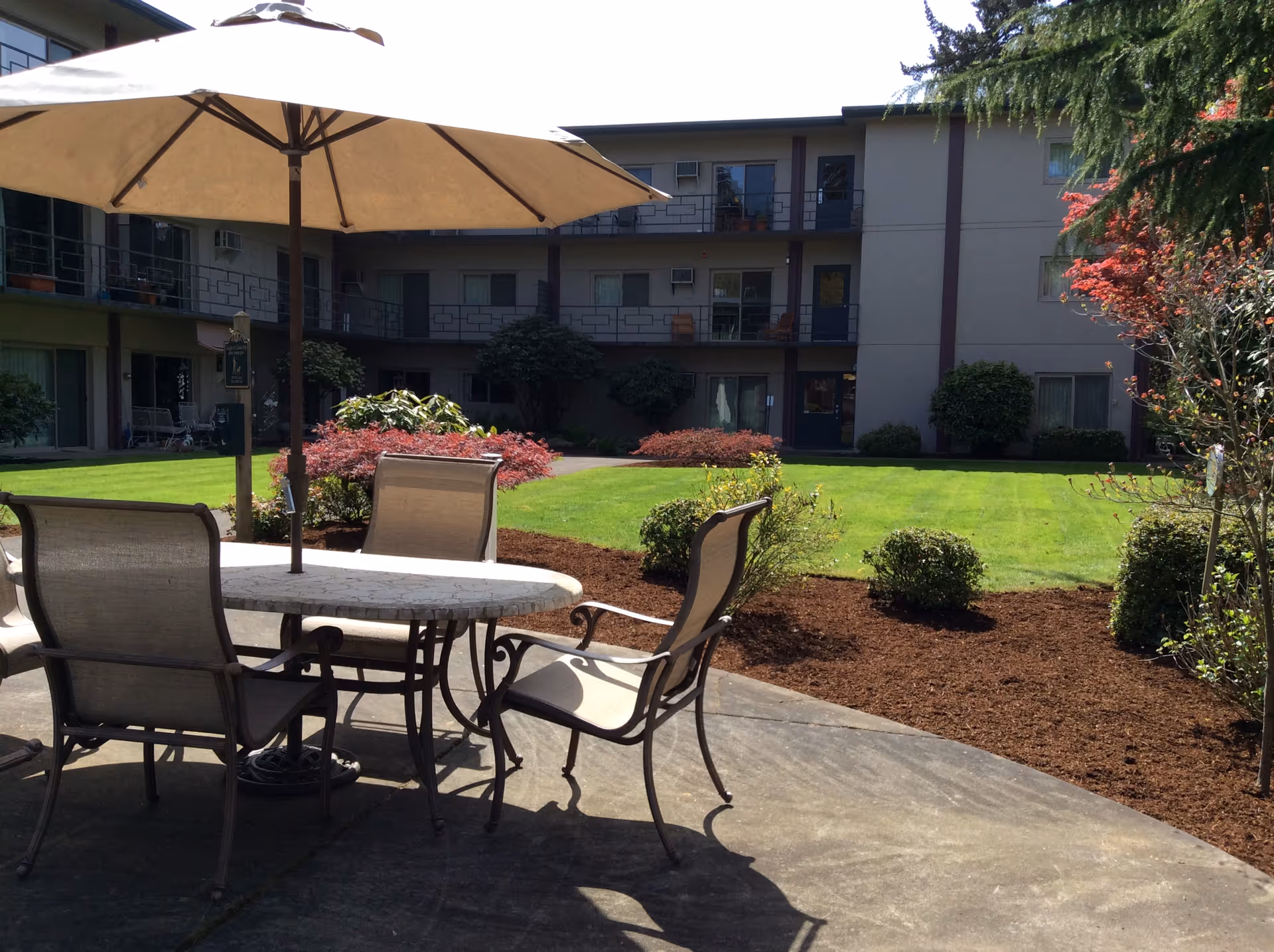 Outdoor courtyard with a patio table, chairs and umbrella overlooking a grassy lawn and multi-story retirement building.