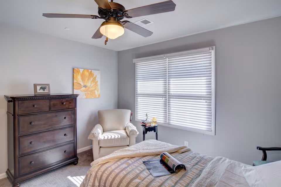 A bright bedroom with a large window covered by white blinds, a beige armchair next to a small black side table with books and a decorative item, a dark wooden dresser with drawers, a ceiling fan with a light, and a bed with a striped comforter and an open magazine on it.