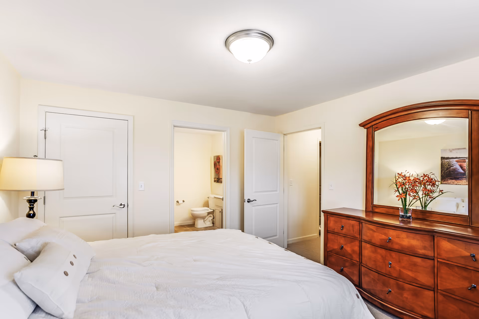 Bright bedroom with white bedding, a bedside lamp, a wooden dresser with mirror and flowers, and an open door showing a bathroom.