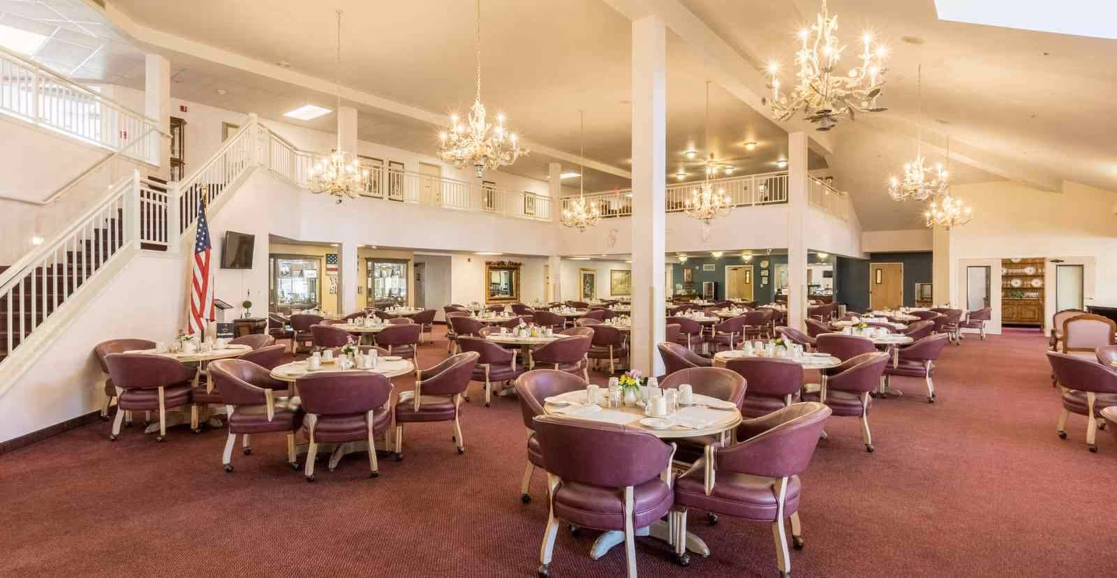 Spacious dining room with multiple round tables set with white tableware and purple cushioned chairs. The room features chandeliers hanging from a high ceiling, a staircase with white railings, an American flag, and a carpeted floor in a warm tone.