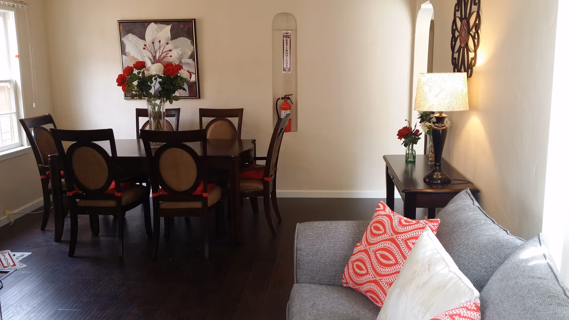 Sunlit dining room with a dark wood table and six chairs, a floral centerpiece and artwork, and a gray sofa with patterned pillows beside a side table lamp.