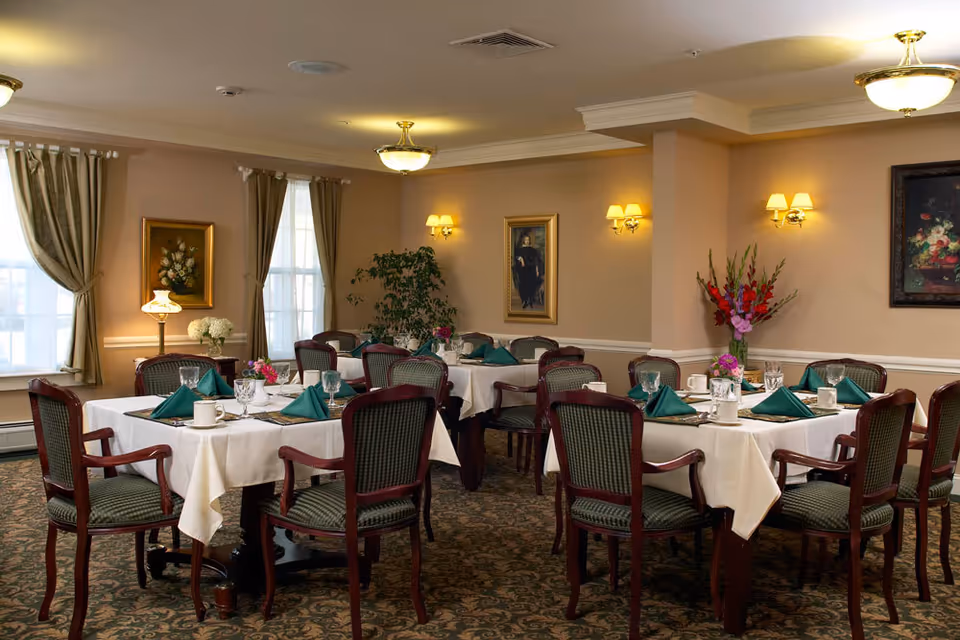 A formal dining room with multiple tables set with white tablecloths, green napkins, place settings and wooden chairs in a senior living facility.