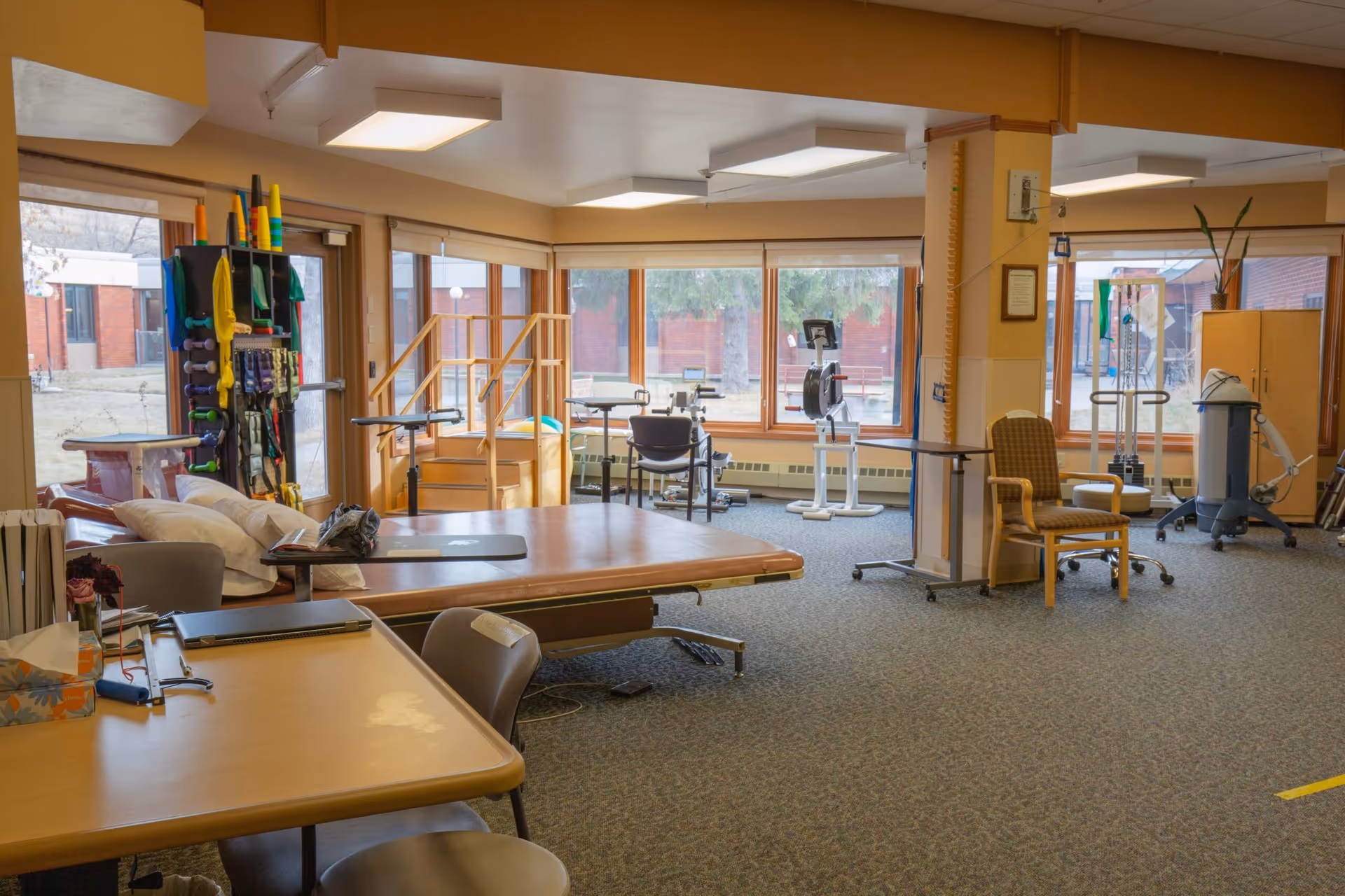 A spacious therapy or rehabilitation room with large windows letting in natural light. The room contains various physical therapy equipment including parallel bars, exercise bikes, and therapy tables. There are chairs and tables arranged around the room, with some therapy tools and supplies visible. The room has a carpeted floor and beige walls.