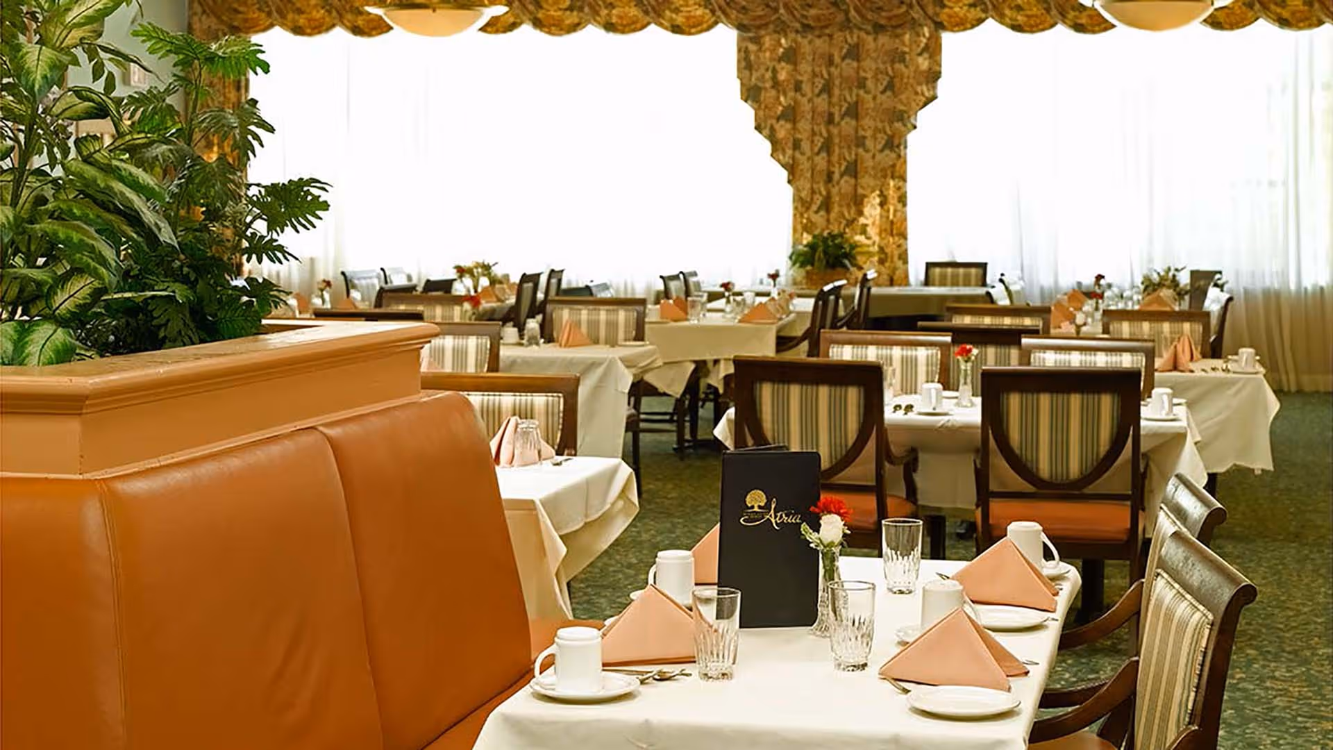 A well-lit dining room with multiple tables covered in white tablecloths, each set with glasses, cups, napkins folded in a triangle shape, and small flower vases. There are cushioned chairs with striped upholstery and a tan leather bench seat. Large windows with sheer curtains and floral drapes allow natural light to fill the room. A menu labeled 'Atria' is placed on one of the tables.