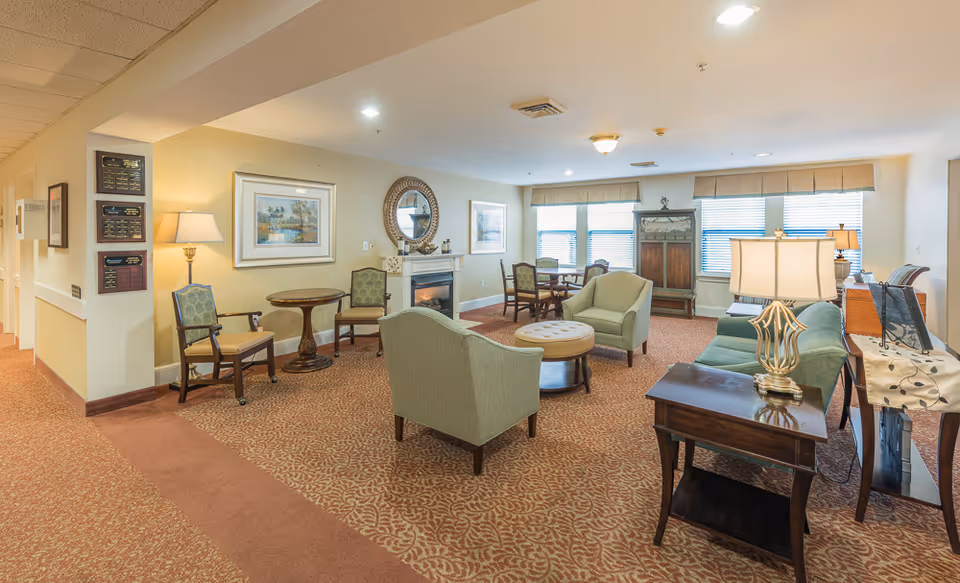 Well-lit communal living room in a senior facility with upholstered chairs and sofas arranged around tables and a fireplace.