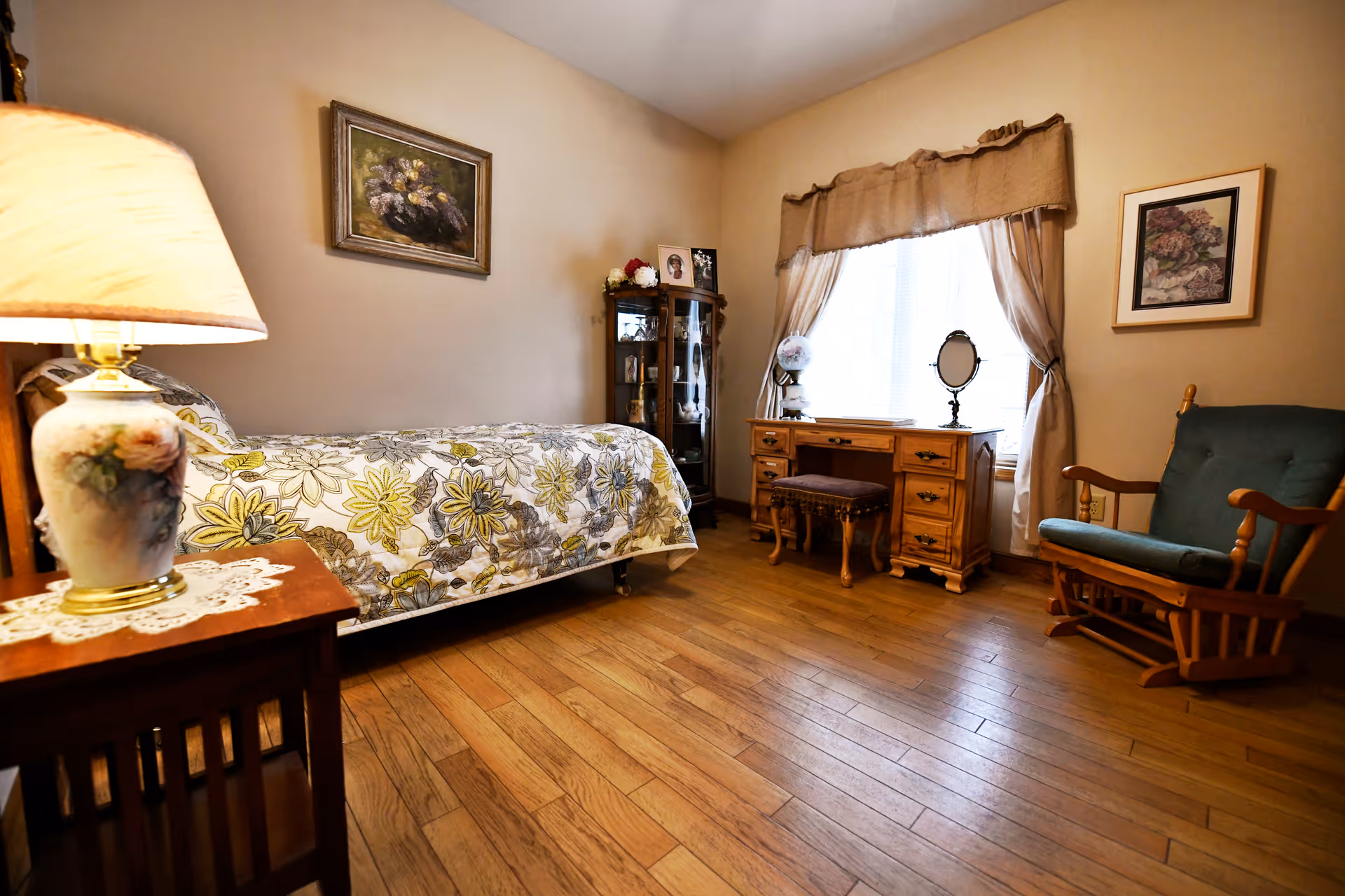 A cozy bedroom with a single bed covered in a floral patterned bedspread, a wooden nightstand with a decorative lamp, a wooden dresser with a mirror and a cushioned stool in front of a window with beige curtains, a wooden rocking chair with green cushions, and framed floral artwork on the walls. The room has wooden flooring and soft beige walls.