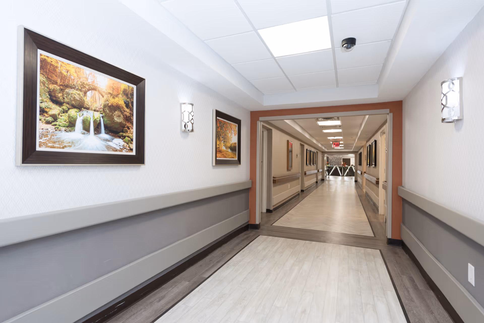 A clean, well-lit hallway in a senior living facility with framed nature photographs on the walls, handrails on both sides, and a light-colored floor leading to another corridor.