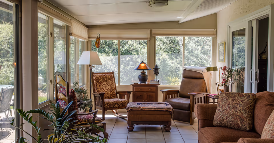 A cozy sunroom with large windows showing green trees outside. The room features a variety of seating including a patterned rocking chair, a brown leather recliner, a cushioned ottoman, and a brown sofa with decorative pillows. There is a wooden side table with a lamp and decorative items, a floor lamp, and several plants adding greenery to the space.