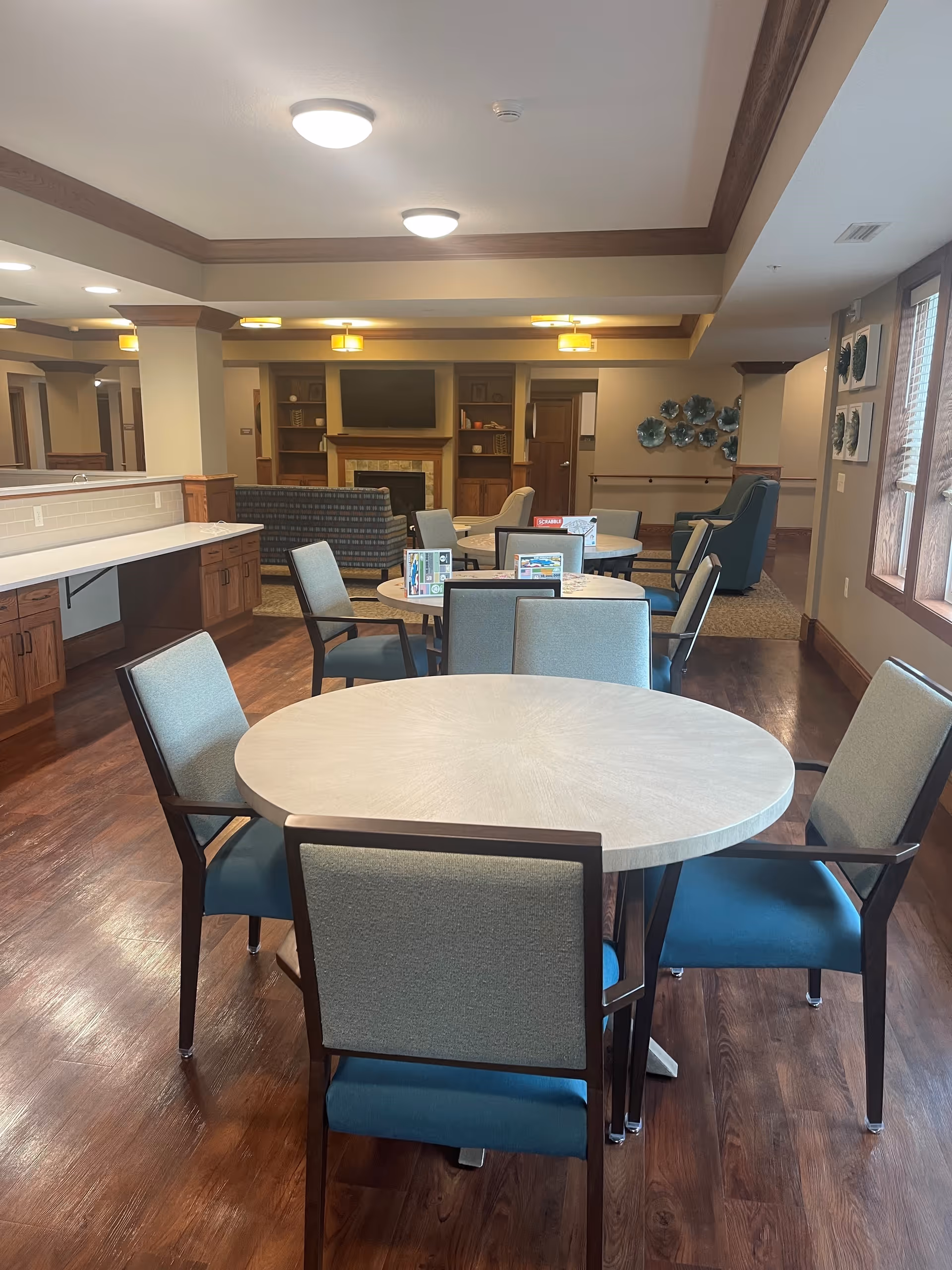 A senior living community common dining area with round tables and chairs, a countertop on the left, and a seating area with a fireplace in the background.