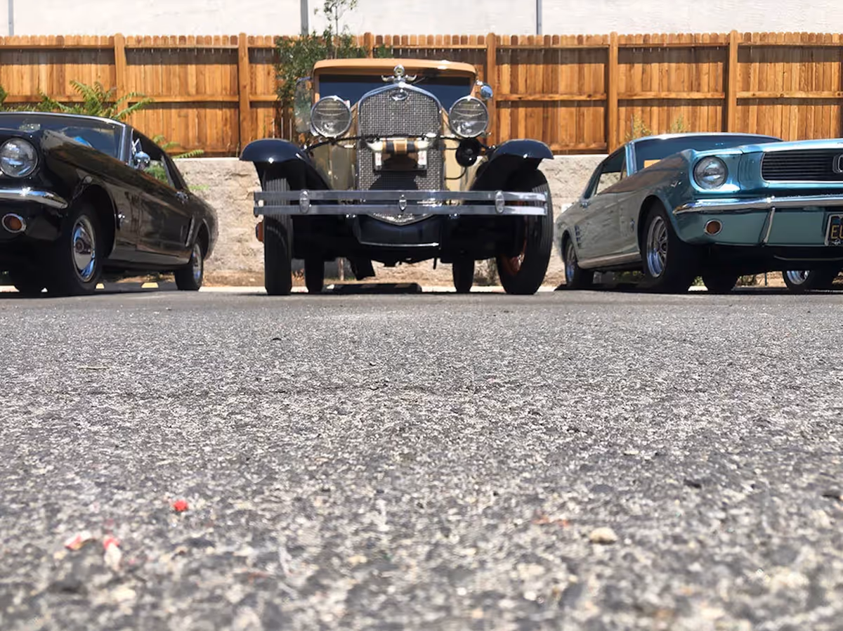Low-angle view of three vintage cars parked side by side on an asphalt surface with a wooden fence and some greenery in the background.