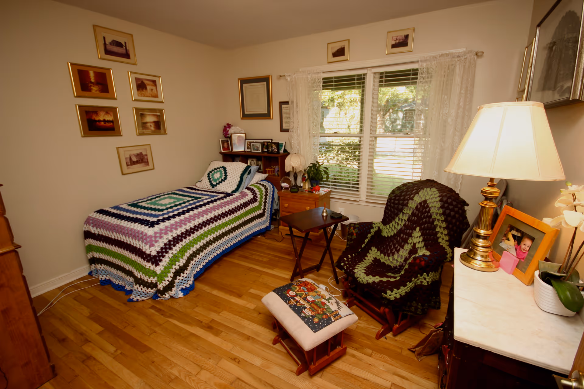 A cozy bedroom with a single bed covered in a colorful crocheted blanket. Next to the bed is a small wooden nightstand with framed photos and a lamp. A cushioned rocking chair with a matching crocheted blanket sits near a window with white lace curtains. A small wooden table and footstool are also present. The room has hardwood floors and several framed pictures on the walls.