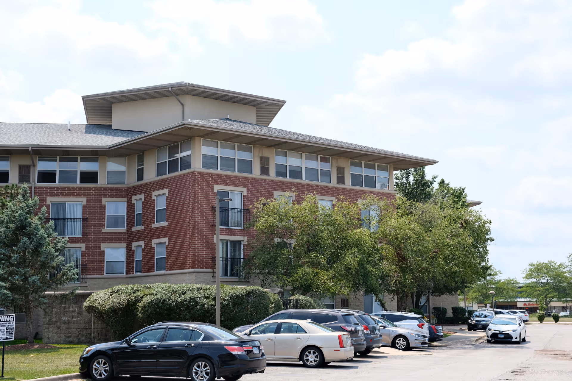 Exterior view of a multi-story senior living facility building with red brick and beige accents, surrounded by trees and parked cars in the parking lot under a partly cloudy sky.