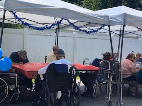 A group of elderly people sitting under white canopy tents outdoors, some in wheelchairs and others in regular chairs, gathered around tables covered with red tablecloths. Blue balloons and blue garlands decorate the tents. A white fence is visible in the background.