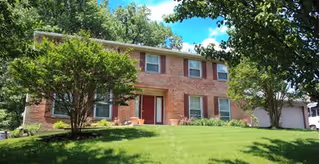 Two-story brick house with a red front door, surrounded by green grass, trees, and shrubs under a blue sky with some clouds.