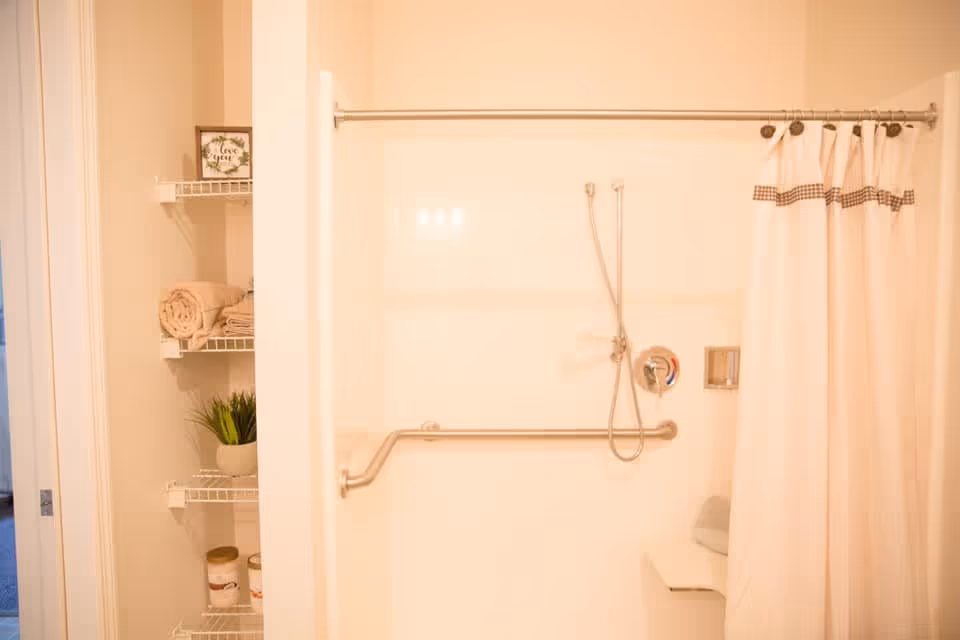 A bathroom shower area with a white shower curtain partially drawn to the right. The shower has a handheld showerhead, a grab bar, and a built-in soap holder. To the left of the shower, there are white wire shelves holding rolled towels, a small potted plant, and some containers. The bathroom has a clean and bright appearance.