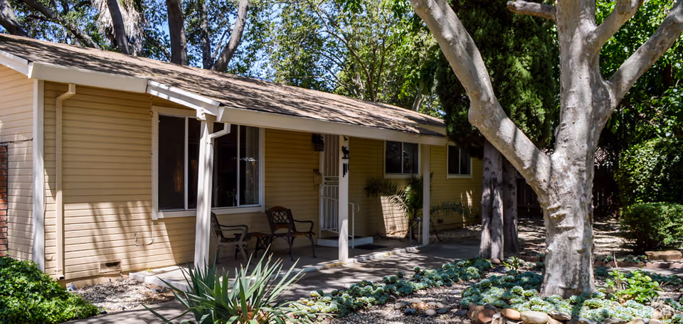Single-story beige building with a small covered porch featuring two chairs. The building is surrounded by trees and landscaped plants, including a large tree in the foreground and various shrubs and ground cover.