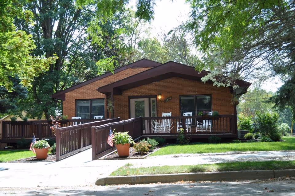 A single-story brick building with a brown roof and a wooden ramp leading to the front porch. The porch has several white rocking chairs and potted plants with small American flags. The building is surrounded by green grass and large trees providing shade.