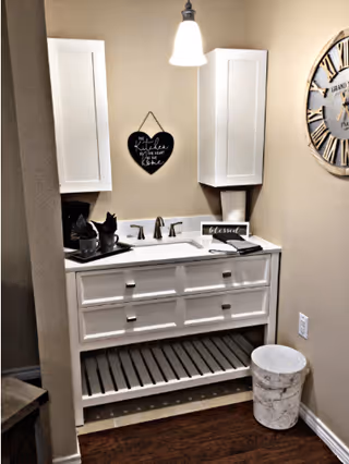 A small white vanity with a sink and faucet is positioned against a beige wall. Above the sink, a white hanging light fixture illuminates a black heart-shaped sign that reads 'The Kitchen is the Heart of the Home.' Two white wall cabinets flank the sign. On the vanity countertop, there is a small decorative item and a sign that says 'blessed.' A large round wall clock is mounted on the right wall. A white trash bin is placed on the floor to the right of the vanity.