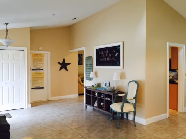 A spacious interior area with beige walls and tiled floor. There is a white door on the left, a black star decoration on the wall, and a dark wooden console table with two lamps and decorative items. A green cushioned chair is placed next to the console table. There are two doorways leading to other rooms, and a hanging light fixture is visible on the ceiling.