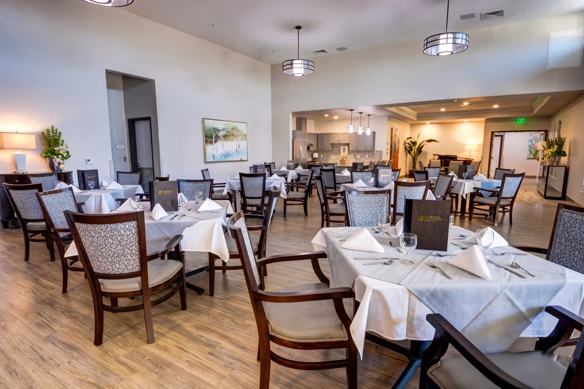 A spacious dining room with multiple tables covered in white tablecloths, each set with folded napkins, glassware, and menus. The room features wooden chairs with patterned upholstery, hardwood flooring, modern pendant lights, and a kitchen area in the background. There are decorative plants and artwork on the walls, creating a welcoming atmosphere.