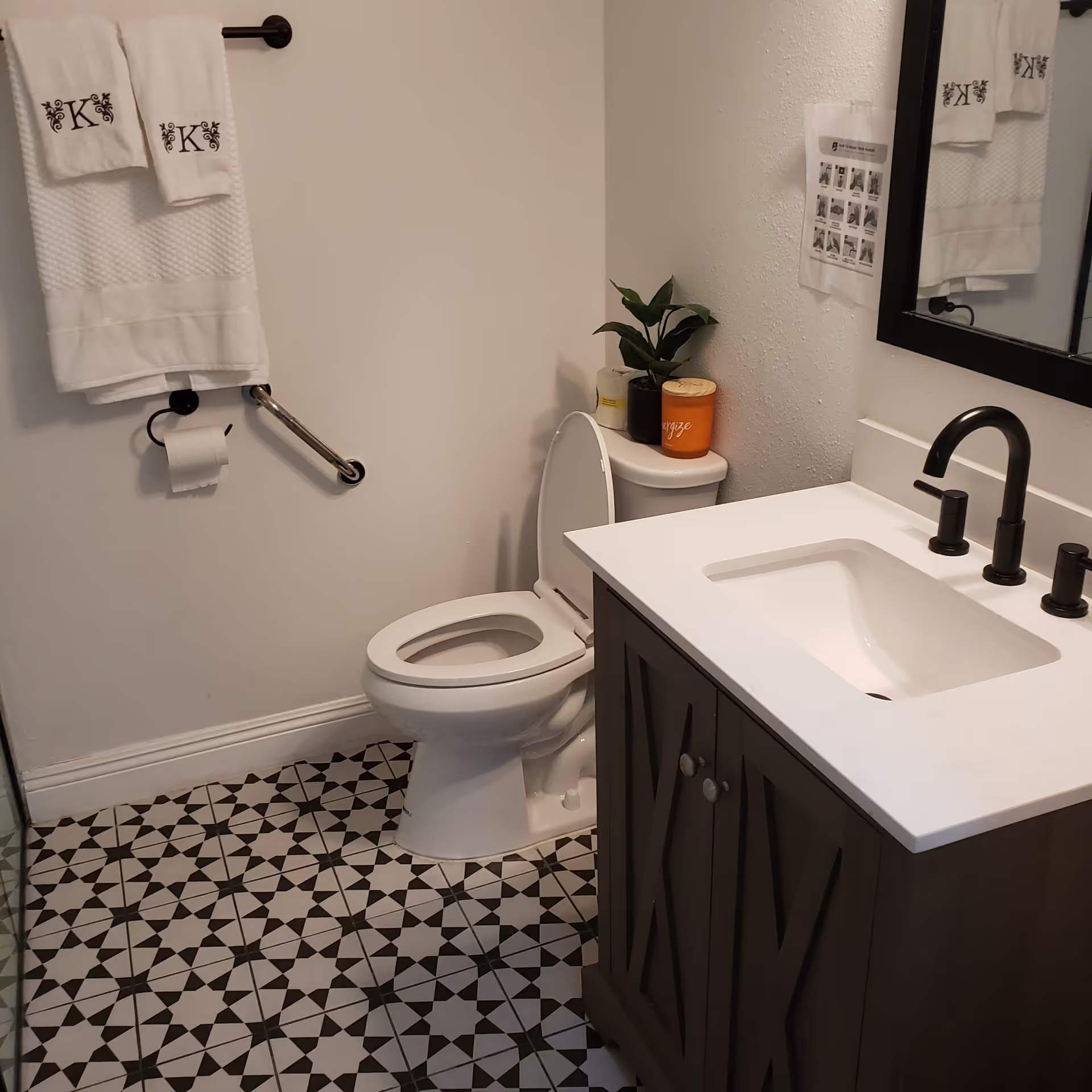 A clean bathroom featuring a white toilet with the lid open, a dark wood vanity with a white countertop and black faucet, a large mirror above the sink, patterned black and white floor tiles, a towel rack with white towels monogrammed with the letter 'K', a toilet paper holder, a grab bar on the wall, and a small plant and candle on the toilet tank.