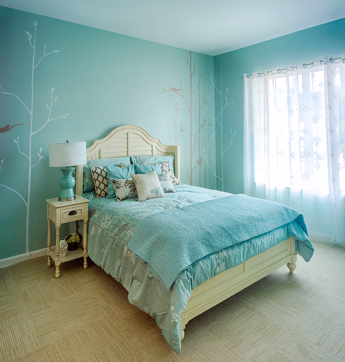 A bedroom with a cream-colored wooden bed frame and a teal bedspread with multiple decorative pillows. The walls are painted teal with a tree and bird mural. There is a small cream-colored nightstand with a teal lamp and decorative items. A window with sheer white curtains allows natural light into the room.