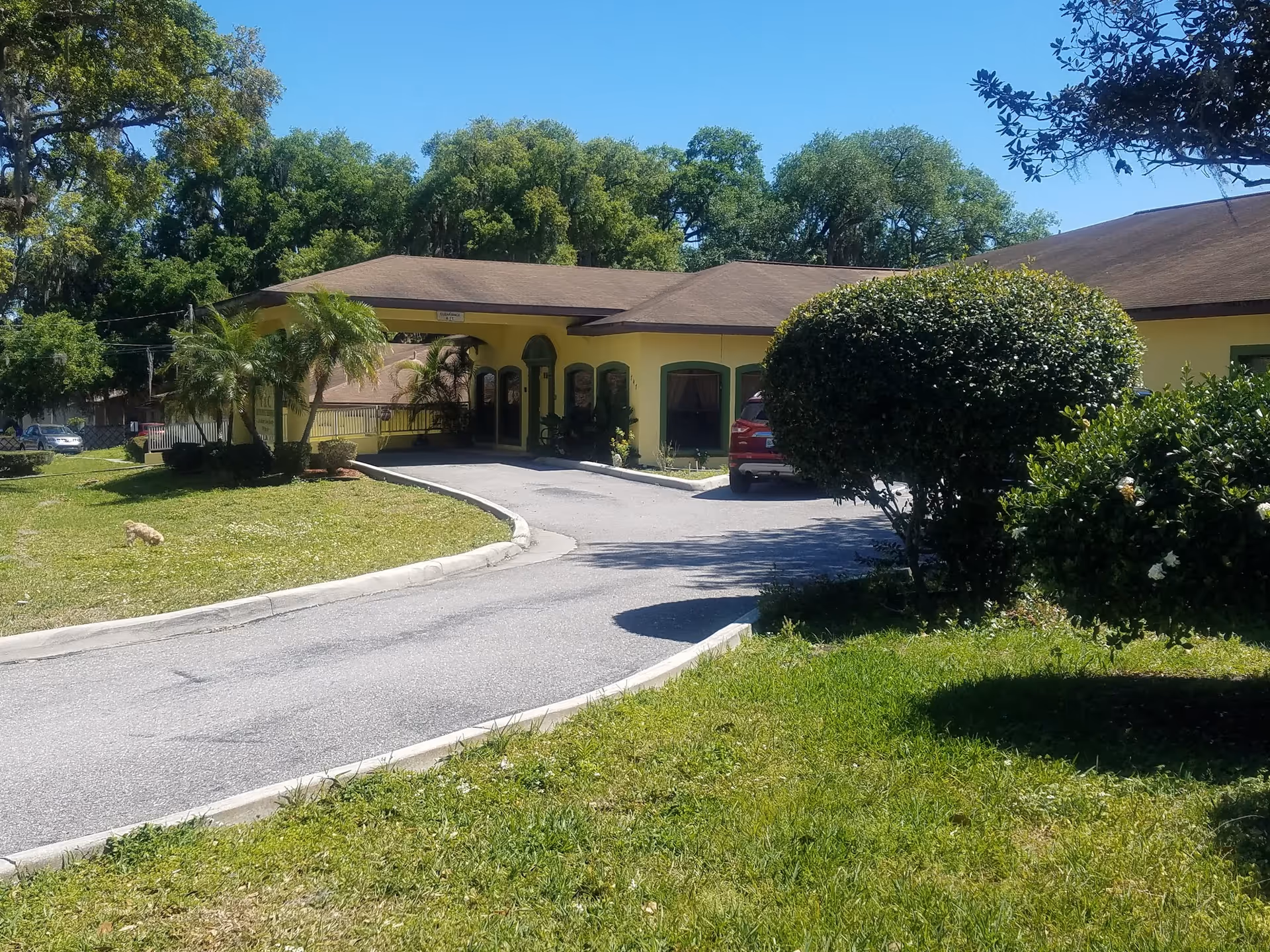 Exterior view of Lakeland Manor Assisted Living Facility showing a single-story yellow building with a brown roof, surrounded by green grass, bushes, and trees under a clear blue sky. A driveway curves up to the entrance where a red car is parked.