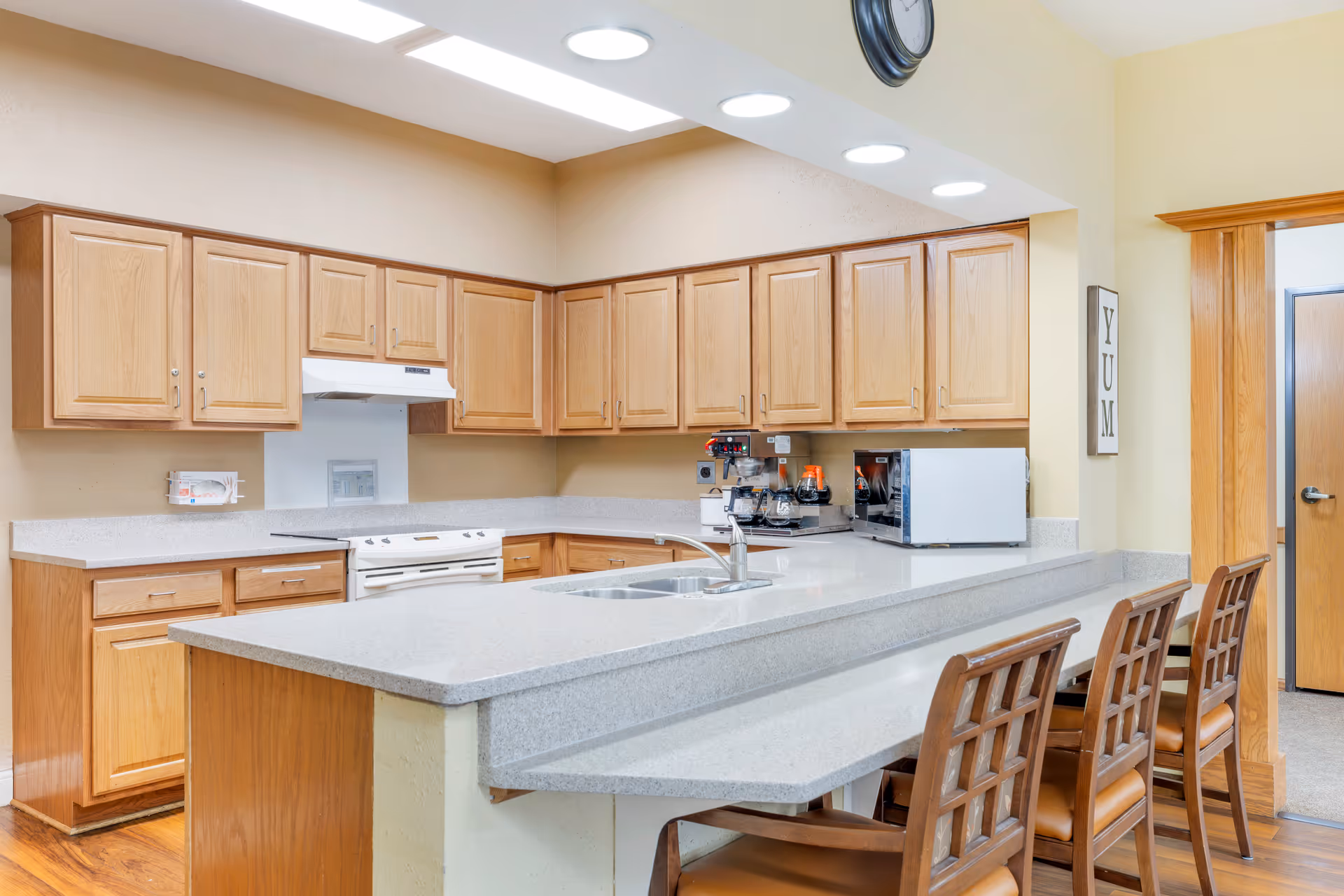 A clean and well-lit kitchen area with wooden cabinets, a white stove, a microwave, a coffee maker, and a double sink. There is a countertop with three wooden chairs for seating. A clock is mounted on the wall above the cabinets, and a sign with the word 'YUM' is hung on the wall near the doorway.