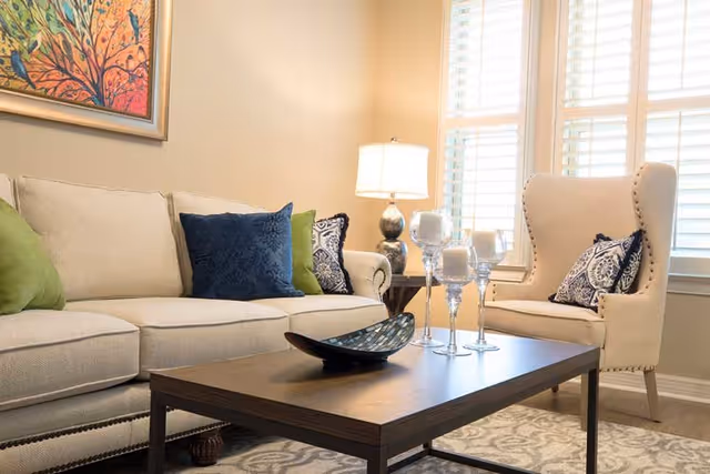 A cozy living room with a beige sofa adorned with green and blue pillows, a wooden coffee table with decorative candles and a bowl, a beige armchair with a patterned pillow, a side table with a lamp, and a colorful painting on the wall.