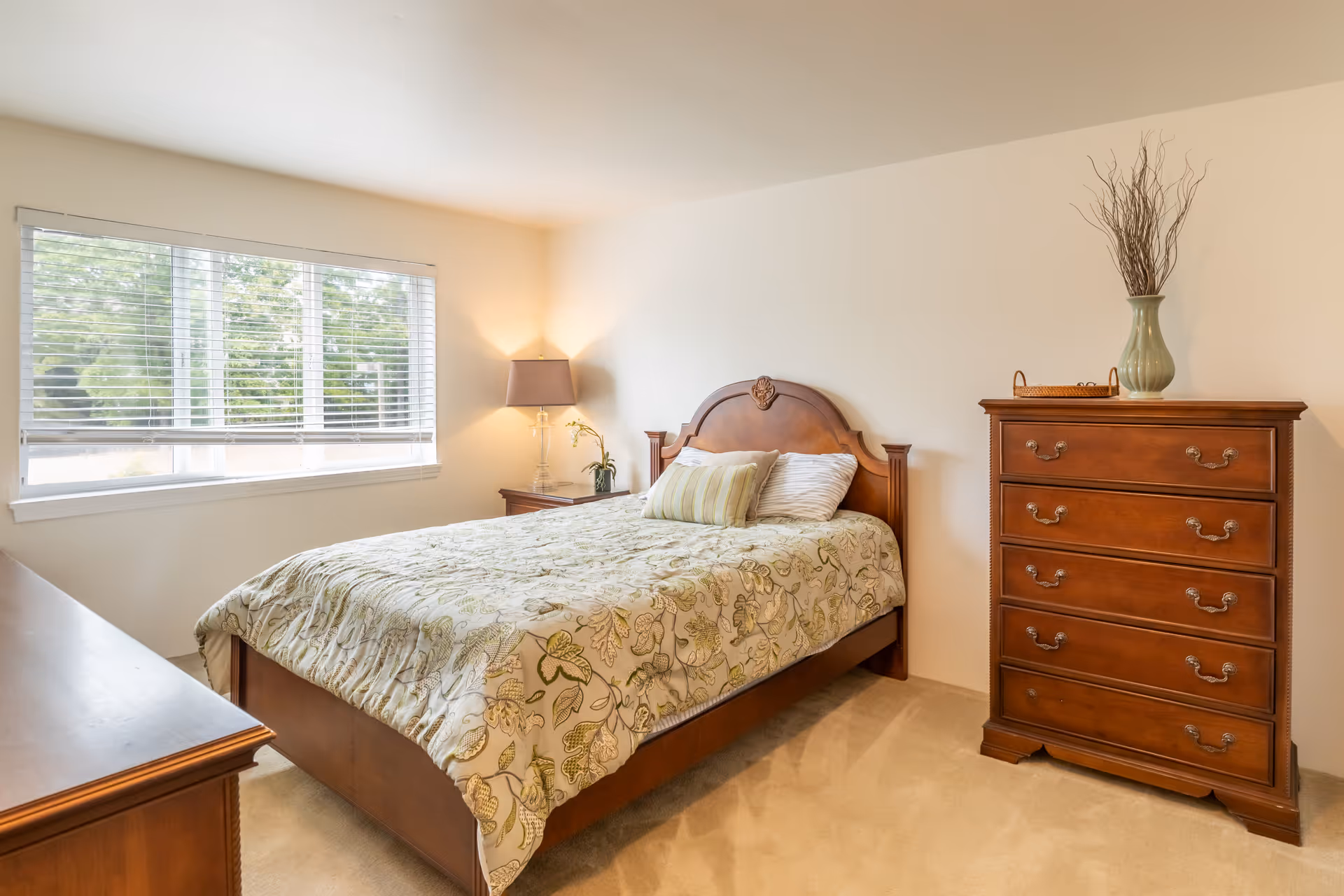 Well-lit bedroom with a wooden bed, floral bedspread, matching dresser and a large window with blinds.