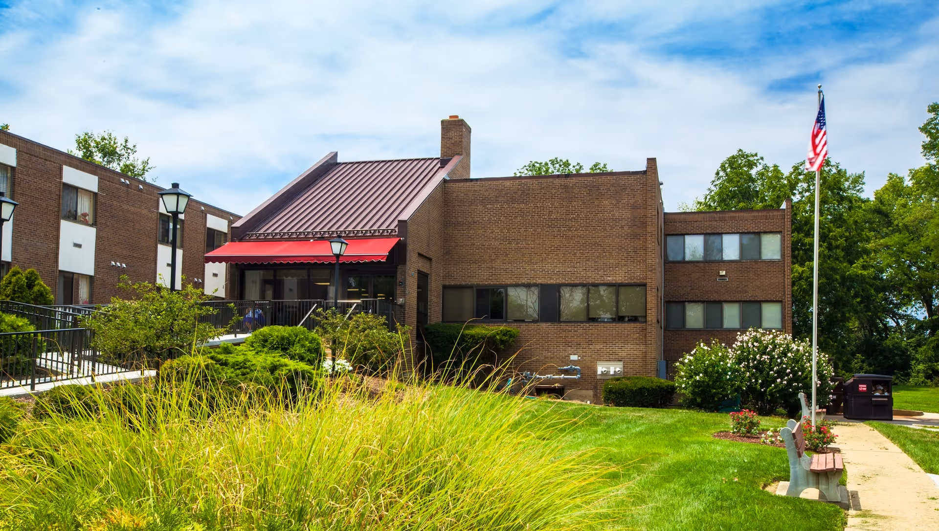 Exterior view of a brick building with a red awning over the entrance, surrounded by green grass, bushes, and trees. There is a flagpole with an American flag and a bench along a sidewalk leading to the entrance.