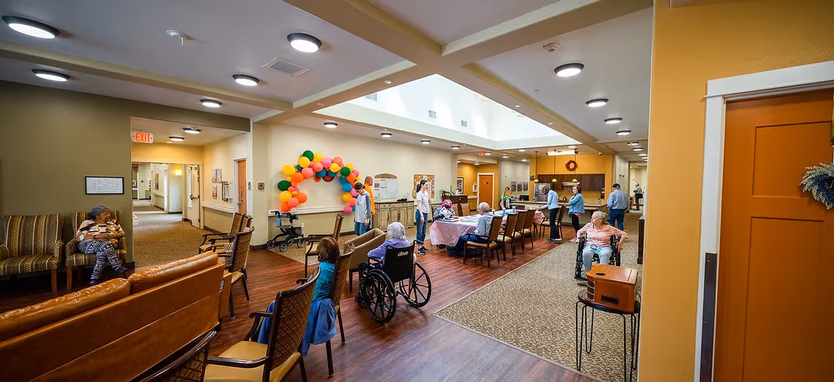 A spacious common area in a senior living facility with several elderly residents, some in wheelchairs, sitting and interacting. The room features a mix of seating including chairs and sofas, a table with people gathered around it, and colorful balloons decorating one wall. The area is well-lit with ceiling lights and natural light from skylights, and has a warm, inviting atmosphere with wood and carpeted flooring.