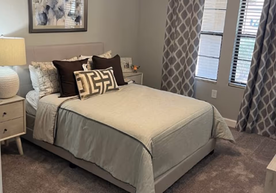 A neatly made bed with decorative pillows, a bedside table and lamp, and patterned curtains in a bedroom.