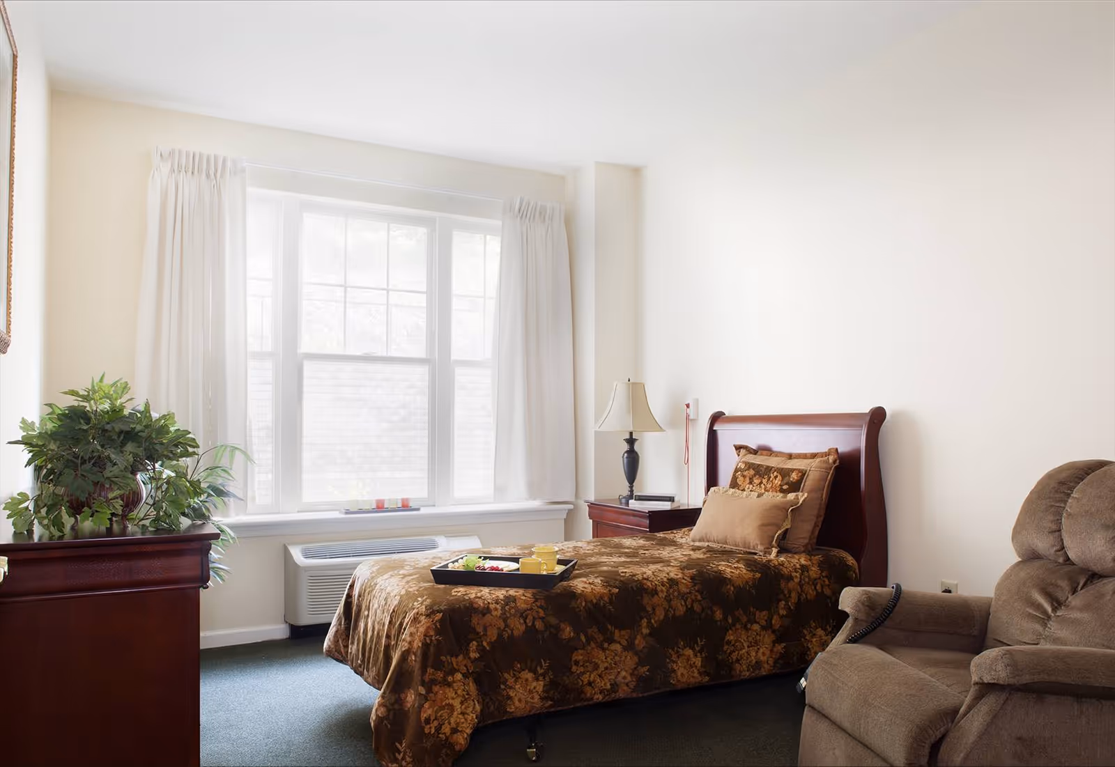 A cozy bedroom with a single bed covered in a brown floral bedspread, a matching pillow, and a tray with breakfast items on the bed. There is a wooden nightstand with a lamp beside the bed, a comfortable brown recliner chair, a wooden dresser with a green plant on top, and a large window with white curtains letting in natural light.