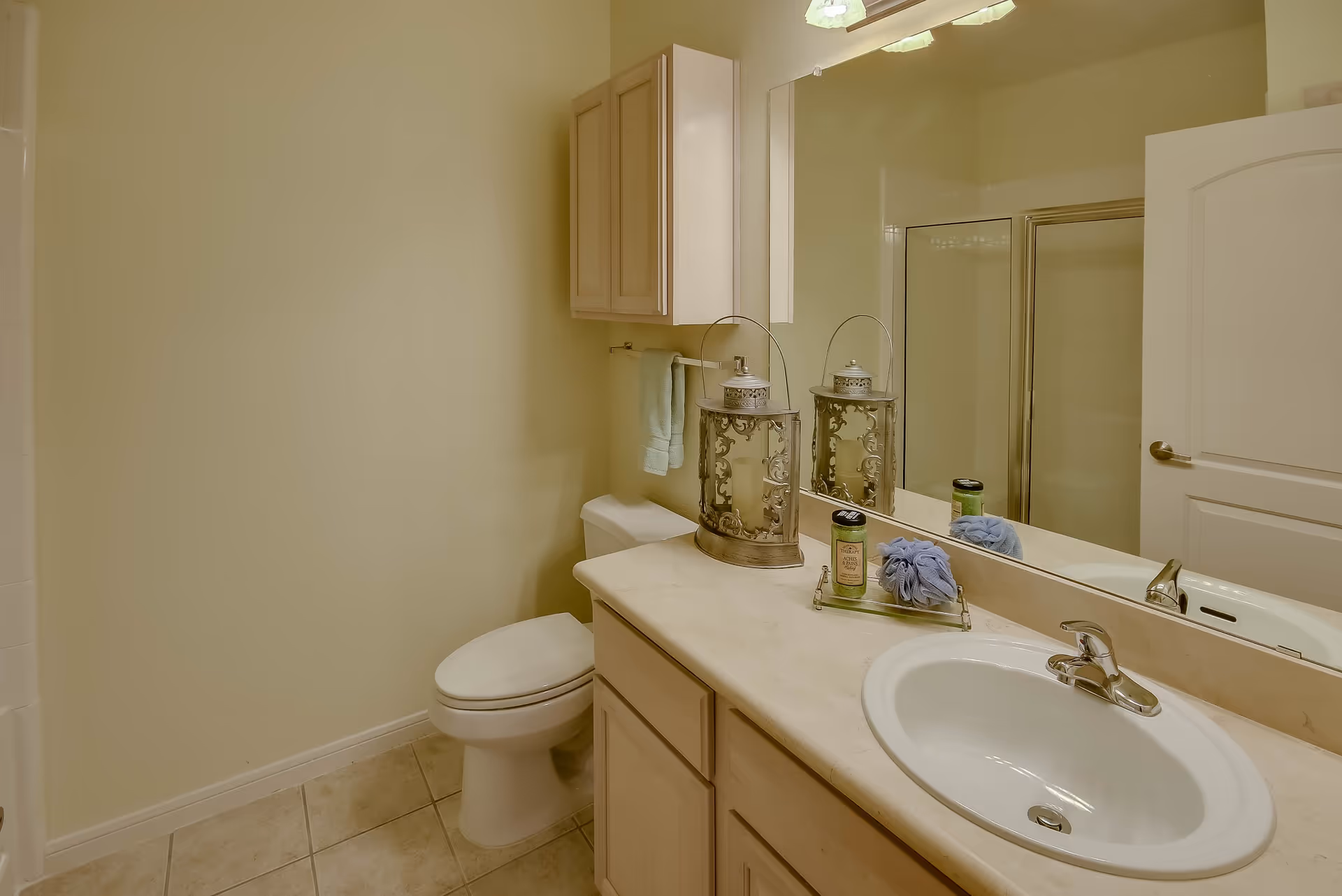 A clean bathroom with a white toilet, a beige countertop with a white sink, a large mirror above the sink, a decorative lantern, a small towel, and a shower with a glass door in the background.