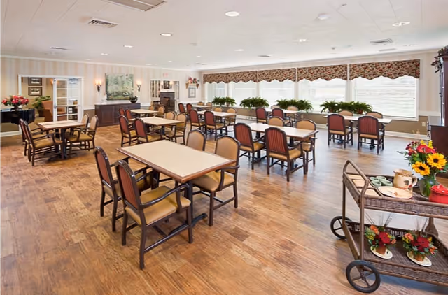 A spacious dining room with multiple tables and chairs arranged neatly. The room has large windows with blinds and floral valances, allowing natural light to fill the space. There are plants on the windowsill and decorative items including flowers on a serving cart in the foreground. The floor is wooden, and the walls are light-colored with framed artwork and sconces.