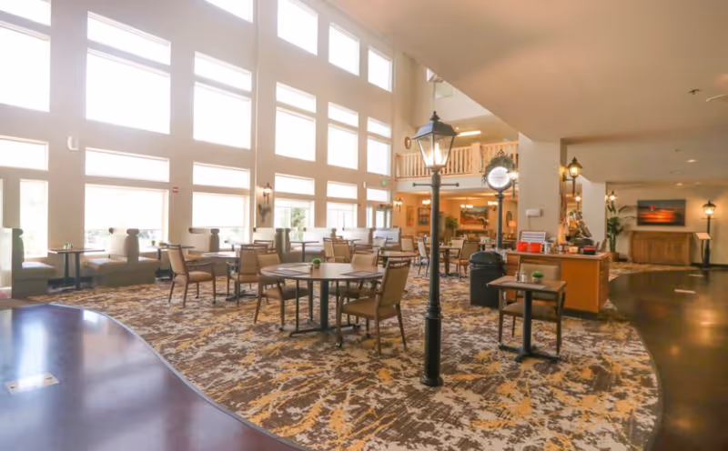 A spacious, well-lit dining area with multiple tables and chairs arranged on a patterned carpet. Large windows allow natural light to fill the room. There are decorative streetlamp-style lights and a clock in the center. The area has a high ceiling and a balcony overlooking the space.