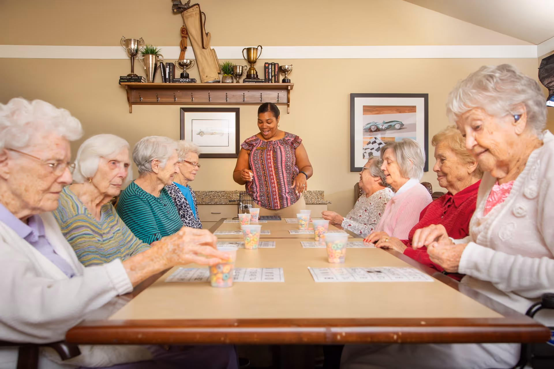 A group of elderly women sitting around a long table playing a game with bingo cards and cups filled with colorful cereal pieces, while a caregiver stands at the head of the table smiling and engaging with them. The room has beige walls with framed pictures and a shelf displaying trophies and books.