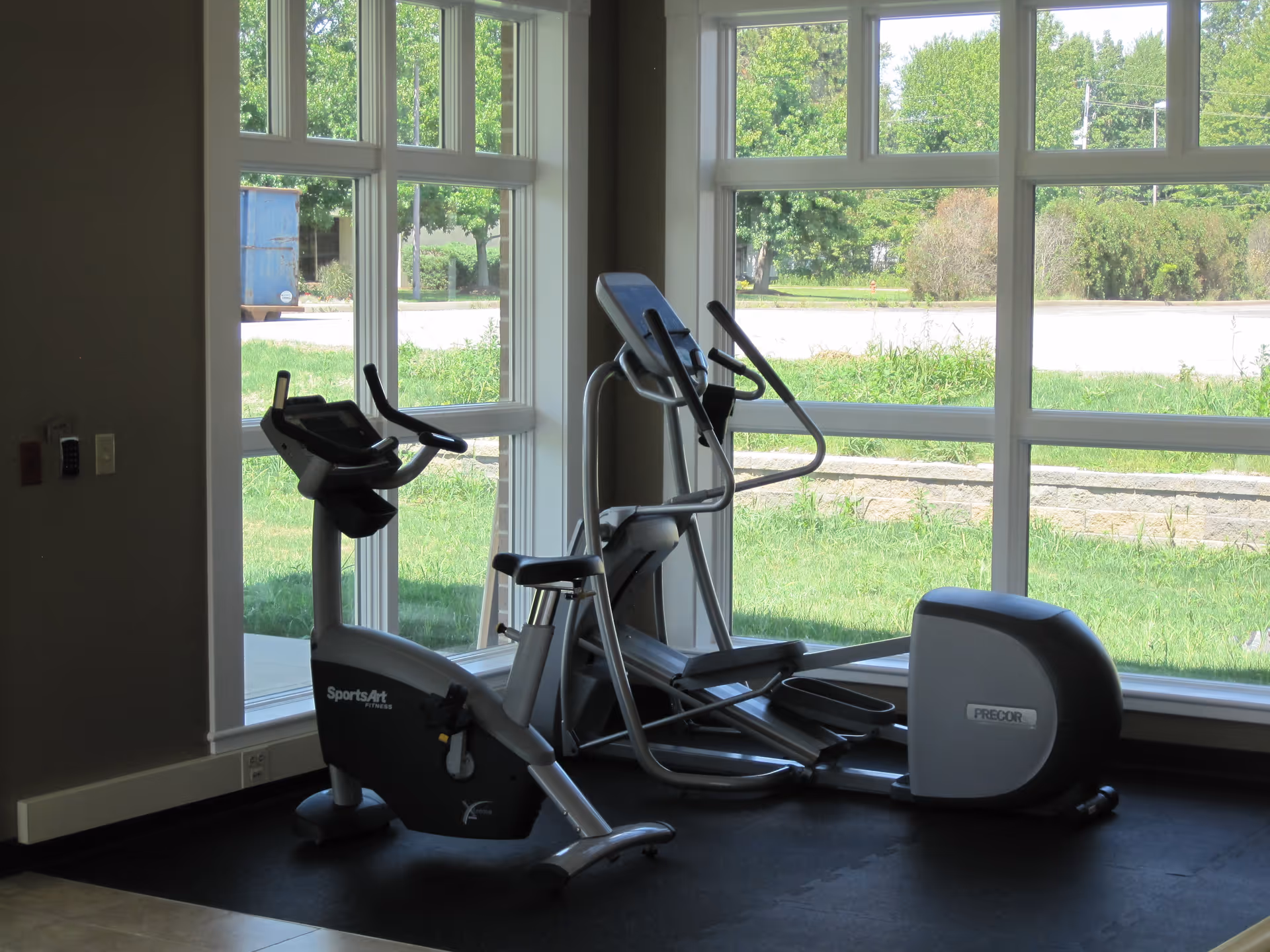 Indoor exercise area with two pieces of fitness equipment, a stationary bike and an elliptical machine, positioned near large windows showing a green outdoor view.