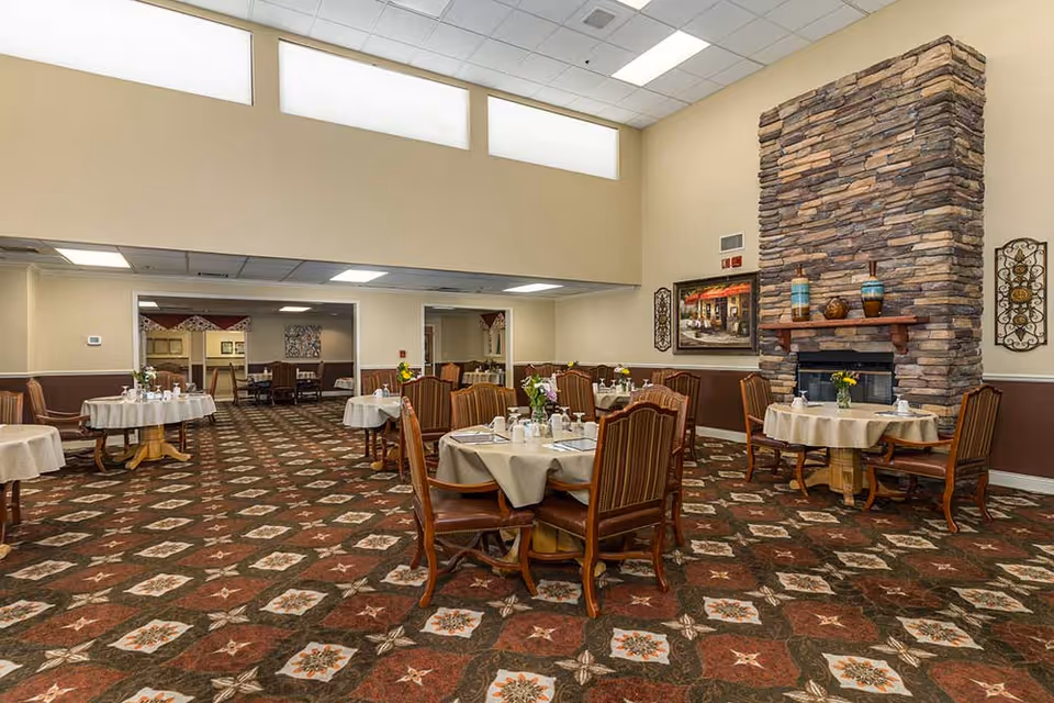Dining room with round tables set for meals, upholstered chairs, patterned carpet, and a stone fireplace.