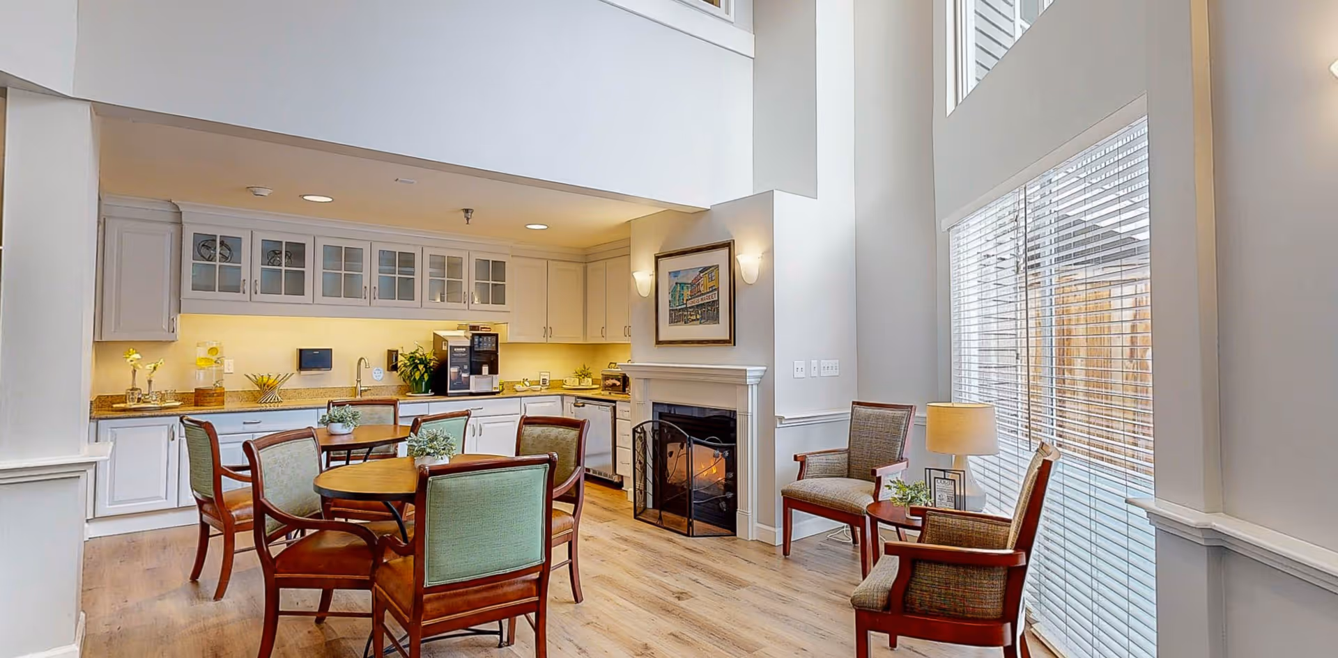 A bright and spacious common area in an assisted living facility featuring a kitchen with white cabinets and countertops, a round wooden table with four chairs, a fireplace with a framed picture above it, and two armchairs with a small side table and lamp near large windows with blinds.