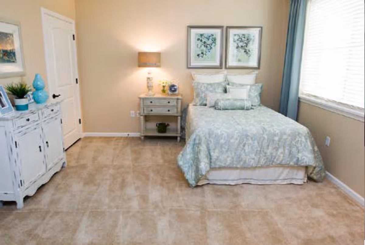 A neatly arranged bedroom with a bed covered in light blue patterned bedding, two framed pictures above the bed, a nightstand with a lamp and decorative items, a white cabinet with plants and a blue vase, beige walls, and a window with blue curtains letting in natural light.