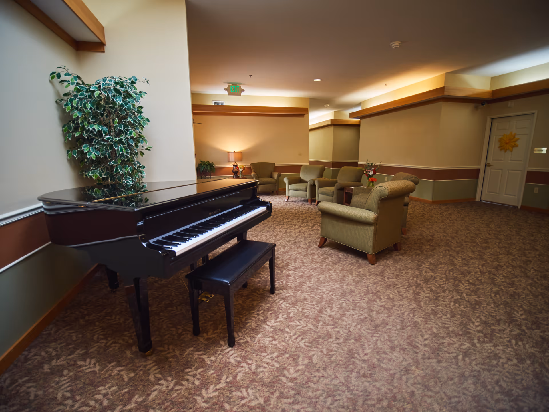 A senior living facility common area with a black grand piano and a matching piano bench on the left side. There is a large potted plant behind the piano. The room has beige walls with wooden trim and patterned carpet flooring. In the background, there are several upholstered armchairs arranged around a small wooden table with a flower vase. A lamp on a side table provides warm lighting, and a door with a sunflower decoration is visible on the right side.
