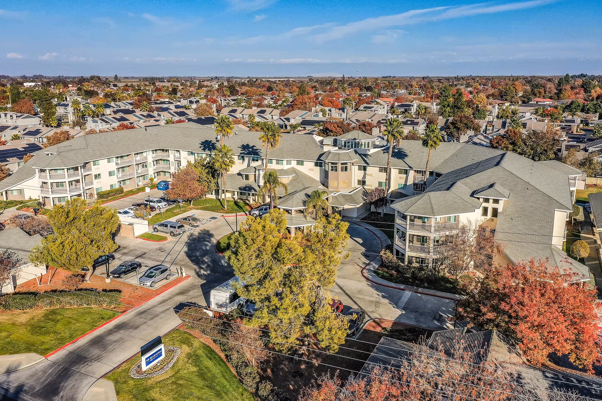 Aerial view of a senior living campus with connected multi-story buildings, parking areas, palm trees, and surrounding neighborhood.