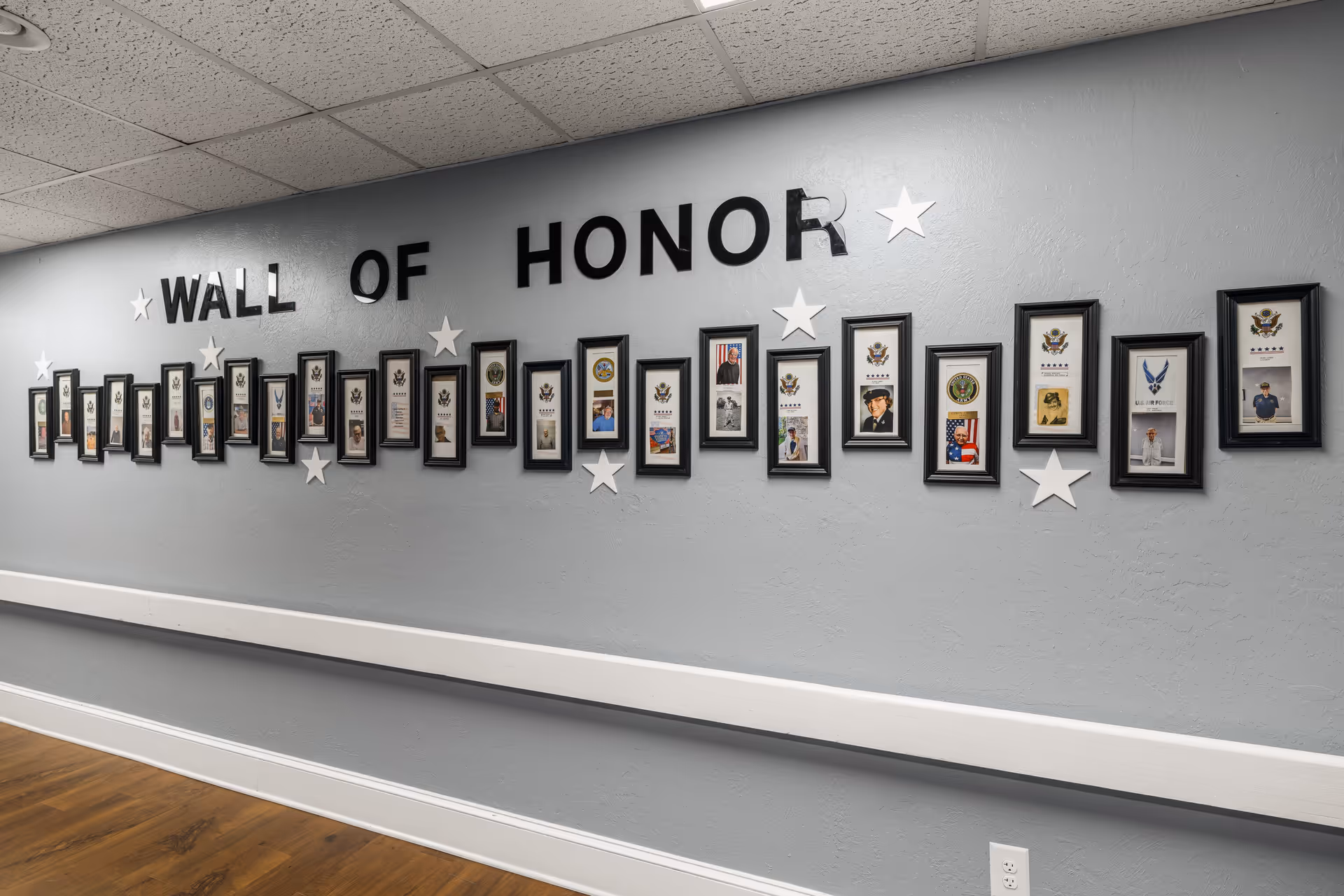A hallway wall painted gray with a display titled 'Wall of Honor' featuring framed photos and military insignias honoring veterans. The wall has black letters and white stars decorating the display. The floor is wood and there is a white chair rail along the wall.