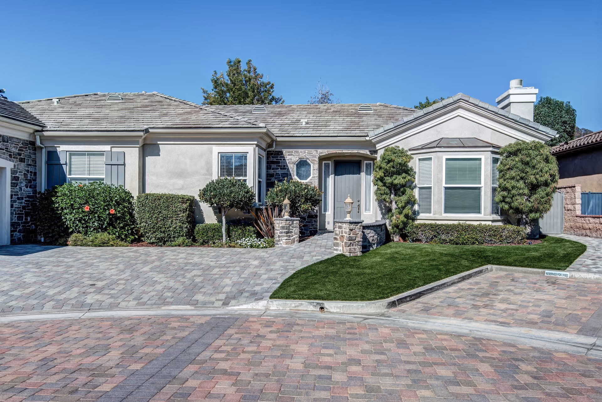 Front exterior view of a single-story residential building with a stone and stucco facade, a tiled roof, manicured bushes, and a paved driveway under a clear blue sky.