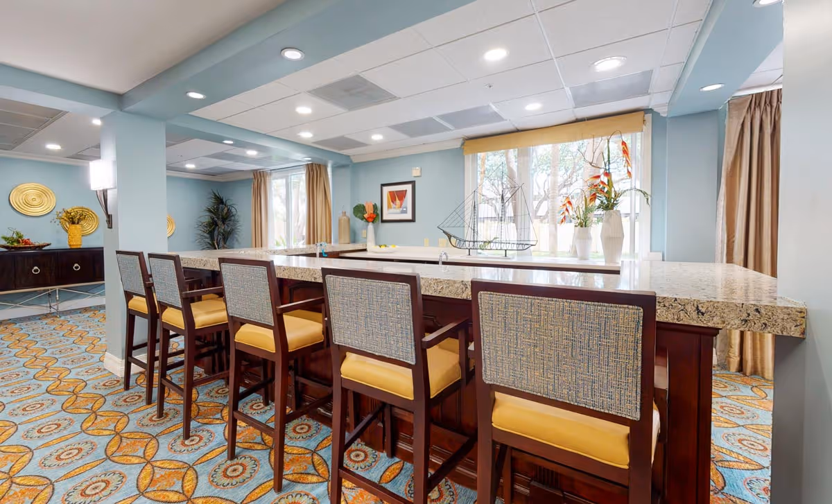 Bright communal dining/lounge area with a long granite-topped bar, five upholstered high-back stools, patterned carpet, and large windows.