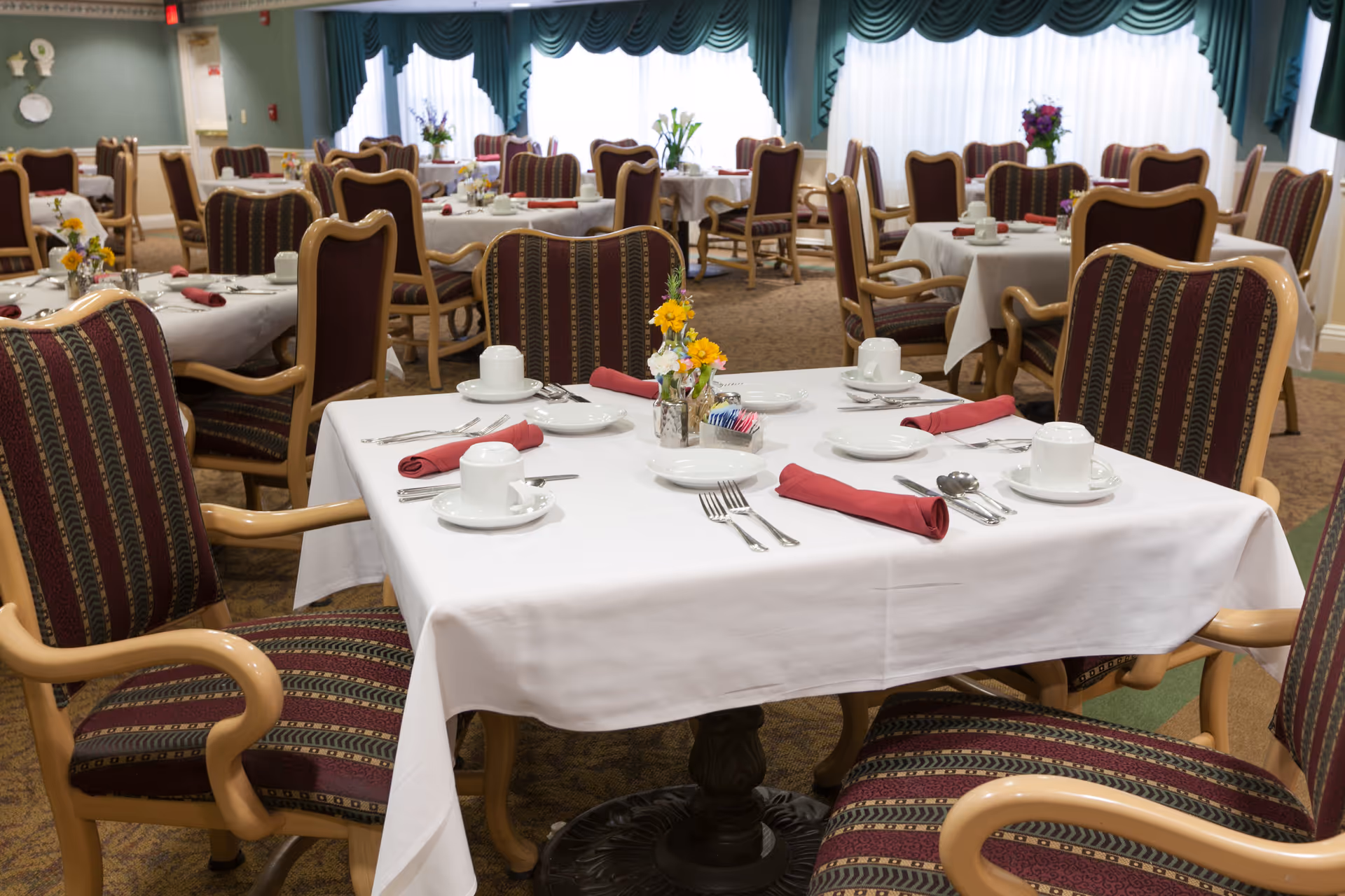 A dining room with multiple tables covered in white tablecloths, each set with white cups, saucers, plates, silverware, and red cloth napkins. The chairs have wooden frames with striped upholstery in shades of burgundy, green, and beige. There are small flower arrangements on the tables and large windows with green valances in the background.