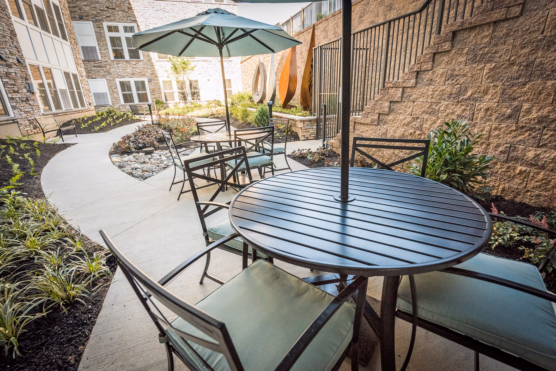 Outdoor patio area at Daylesford Crossing with round metal tables and chairs featuring green cushions. One table has a large green umbrella. The patio is surrounded by a curved concrete walkway, landscaped plants, and a stone wall with decorative metal sculptures.