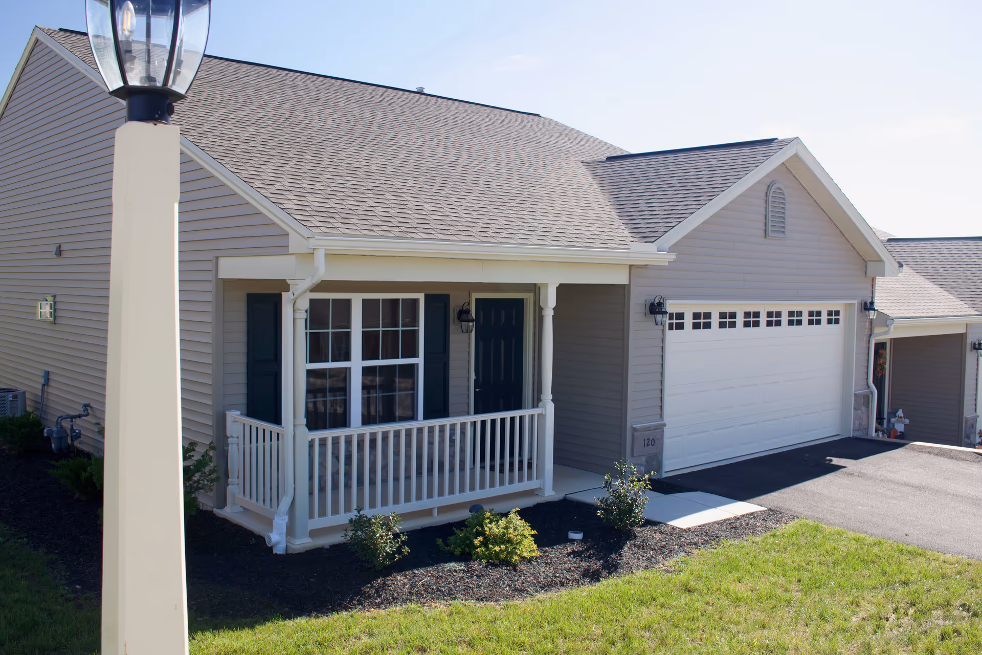 Exterior view of a single-story residential unit with beige siding, a small covered porch with white railing, a black front door, and a white garage door. There is a lamp post in the foreground and some landscaping with small bushes and grass.